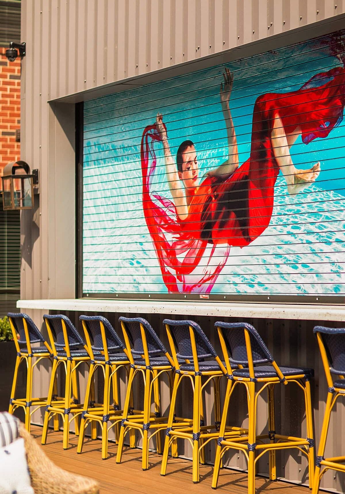 A mural of a woman in a red dress underwater decorates a wall above a row of yellow and blue barstools at an outdoor seating area.