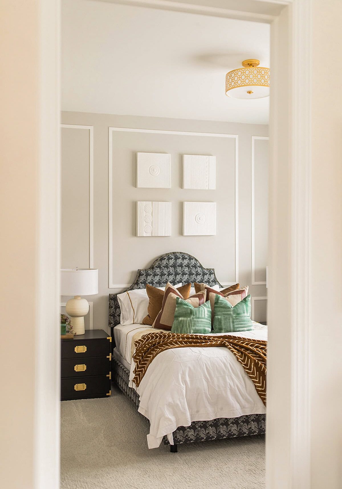 A neatly made bed with patterned pillows and a brown blanket is centered in a modern bedroom with white walls and minimalist wall art.