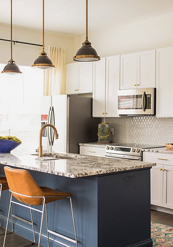Modern kitchen with white cabinets, a blue island, marble countertop, three pendant lights, brown chairs, stainless steel appliances, and a large window with beige curtains.