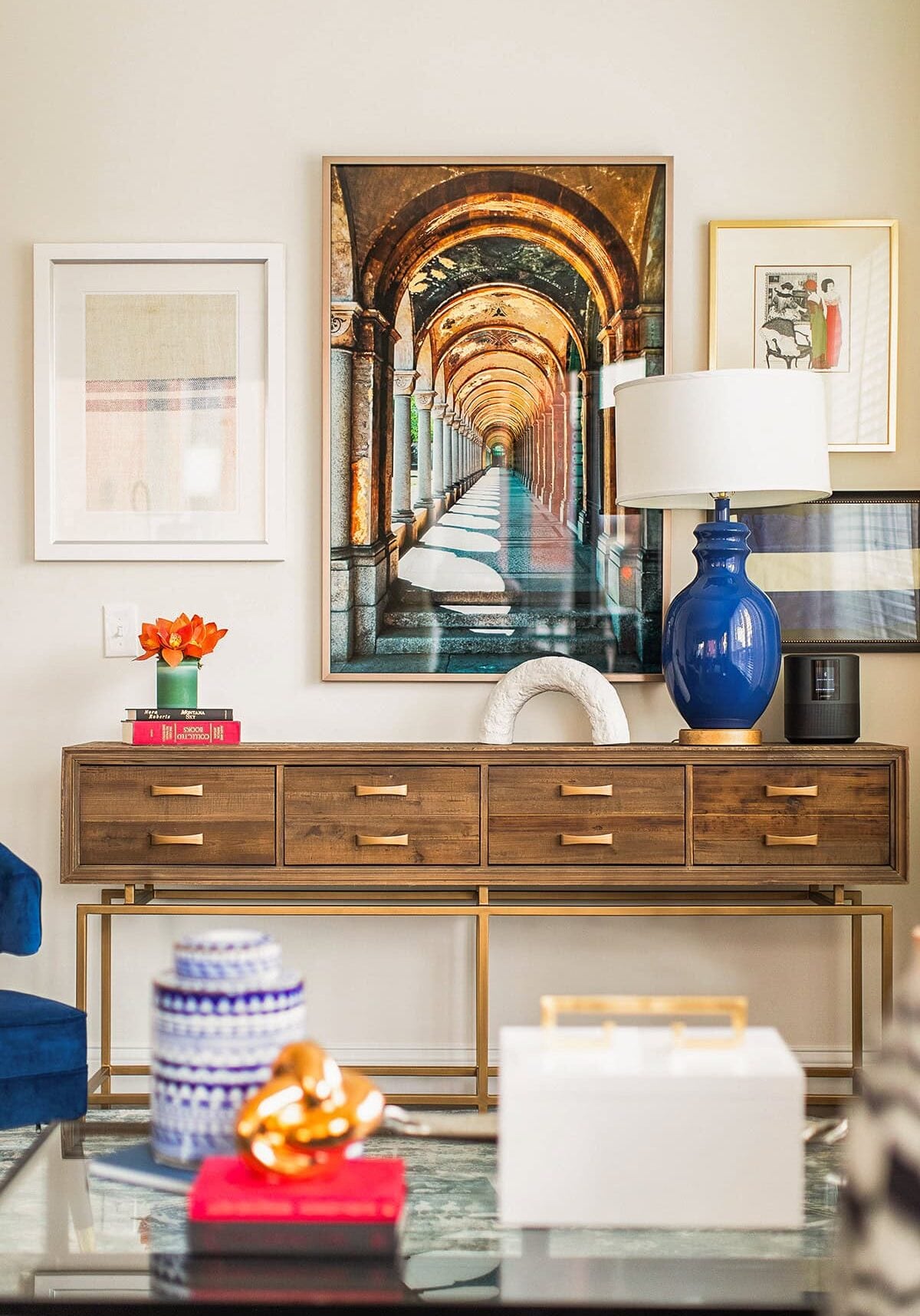 A mid-century modern console table with brass accents sits against a wall, topped with books, a blue lamp, and decor. Framed artwork, including an arched hallway photo, hangs above.