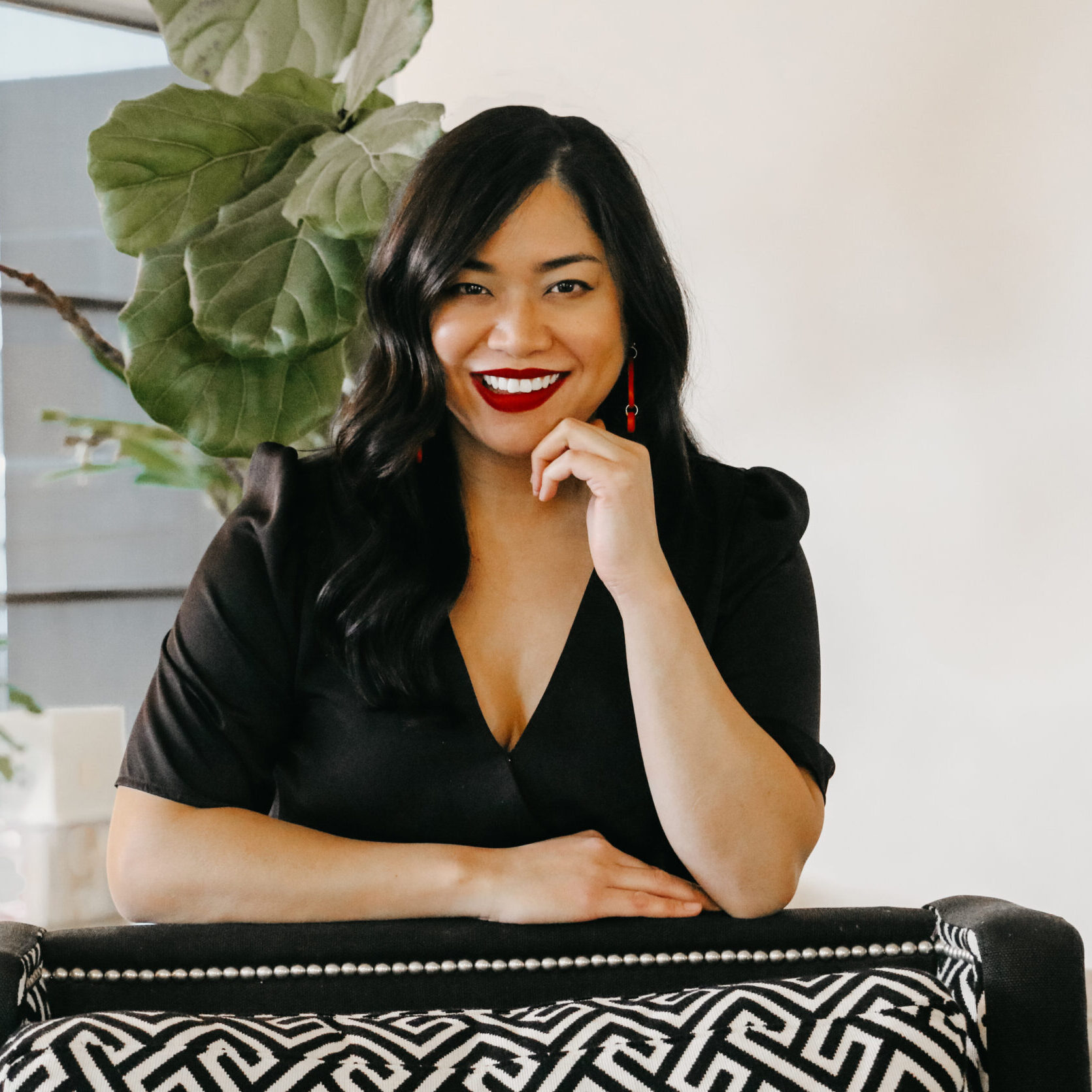 A woman with long black hair and red lipstick sits in a black and white chair, smiling at the camera. Styled by Crimson Design Group, the space features a large green plant in the background.