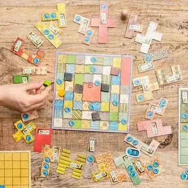 A person's hand moves a piece on a colorful patchwork-themed board game, designed with Crimson Design Group flair, as various game pieces and boards are spread out on a wooden table.