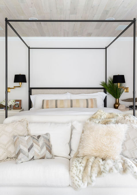 A modern bedroom with a canopy bed, white sofa with decorative pillows in front, neutral walls, wood ceiling, and natural light from large windows with curtains.