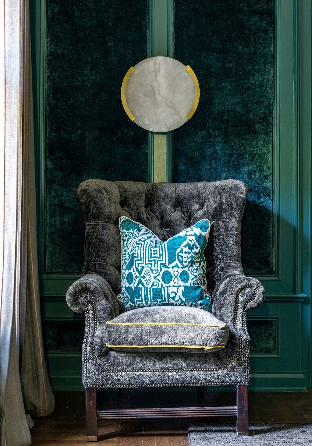 A gray velvet armchair with a blue patterned throw pillow sits against teal paneled walls; a round wall decoration hangs above it.