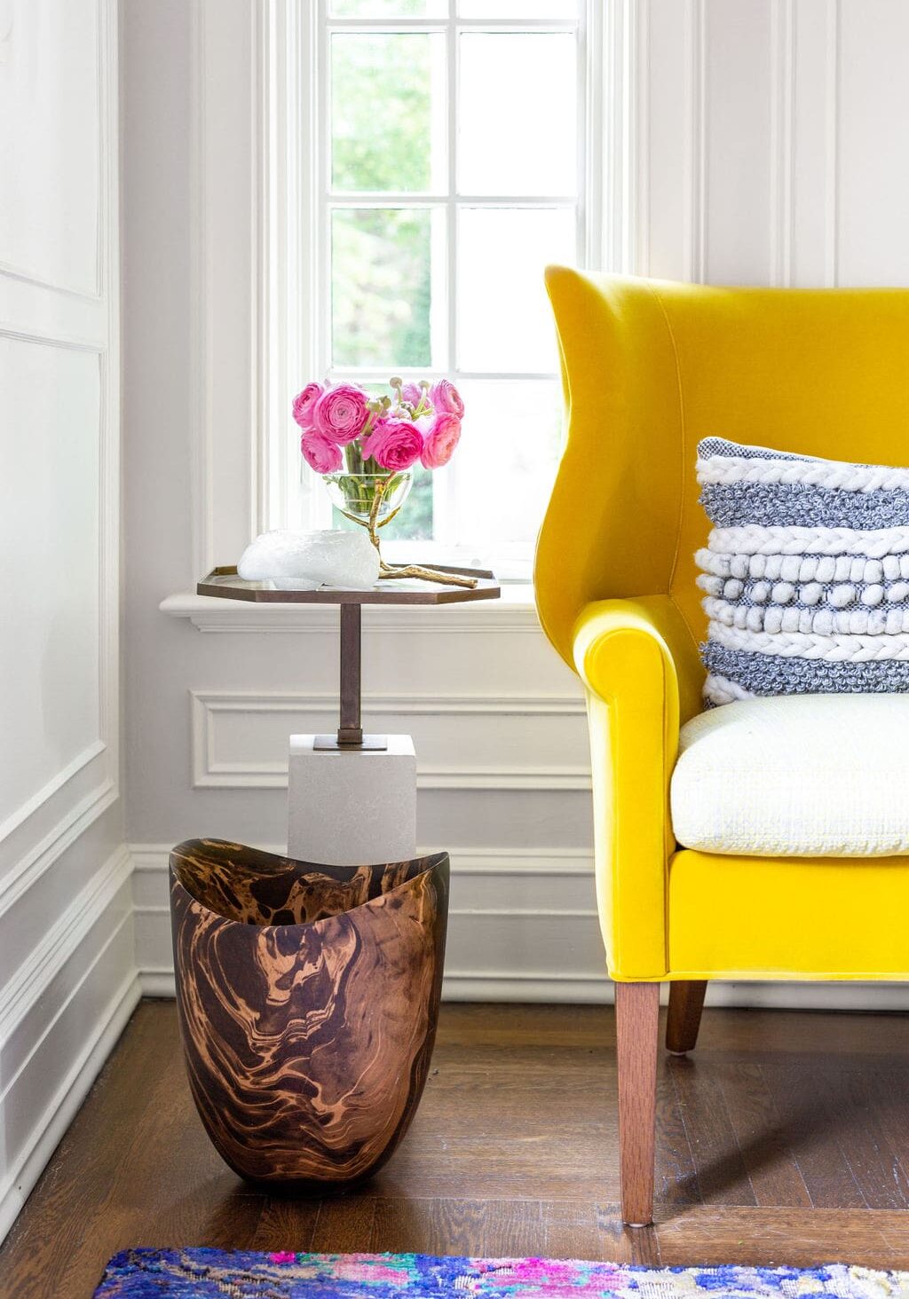 A yellow armchair with textured pillows sits beside a side table holding pink flowers in a vase, next to a wood and marble decorative piece, in a bright room with a window.