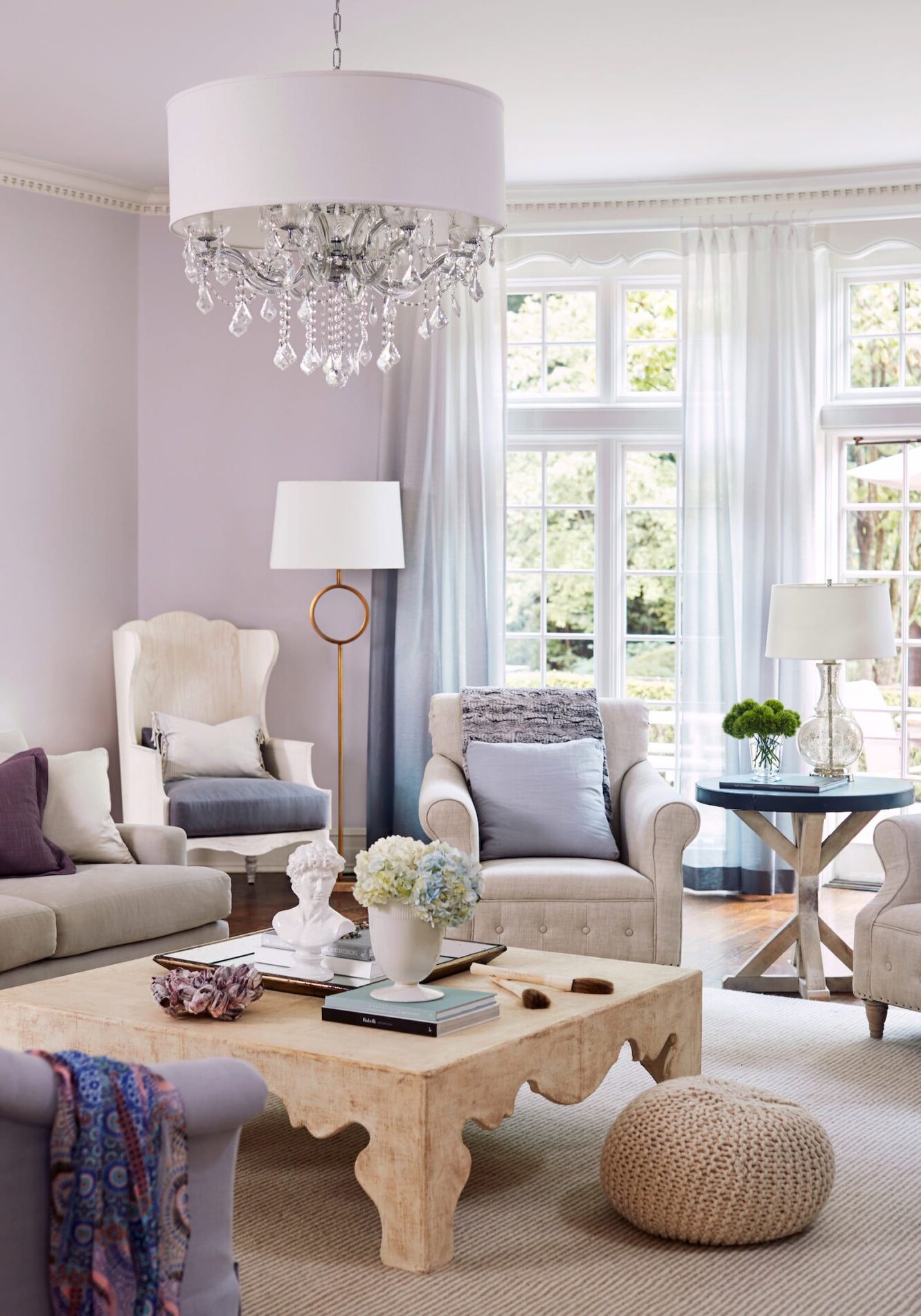 Bright living room with neutral-toned furniture, a wooden coffee table, a crystal chandelier, large windows, and decorative accents like flowers and books.