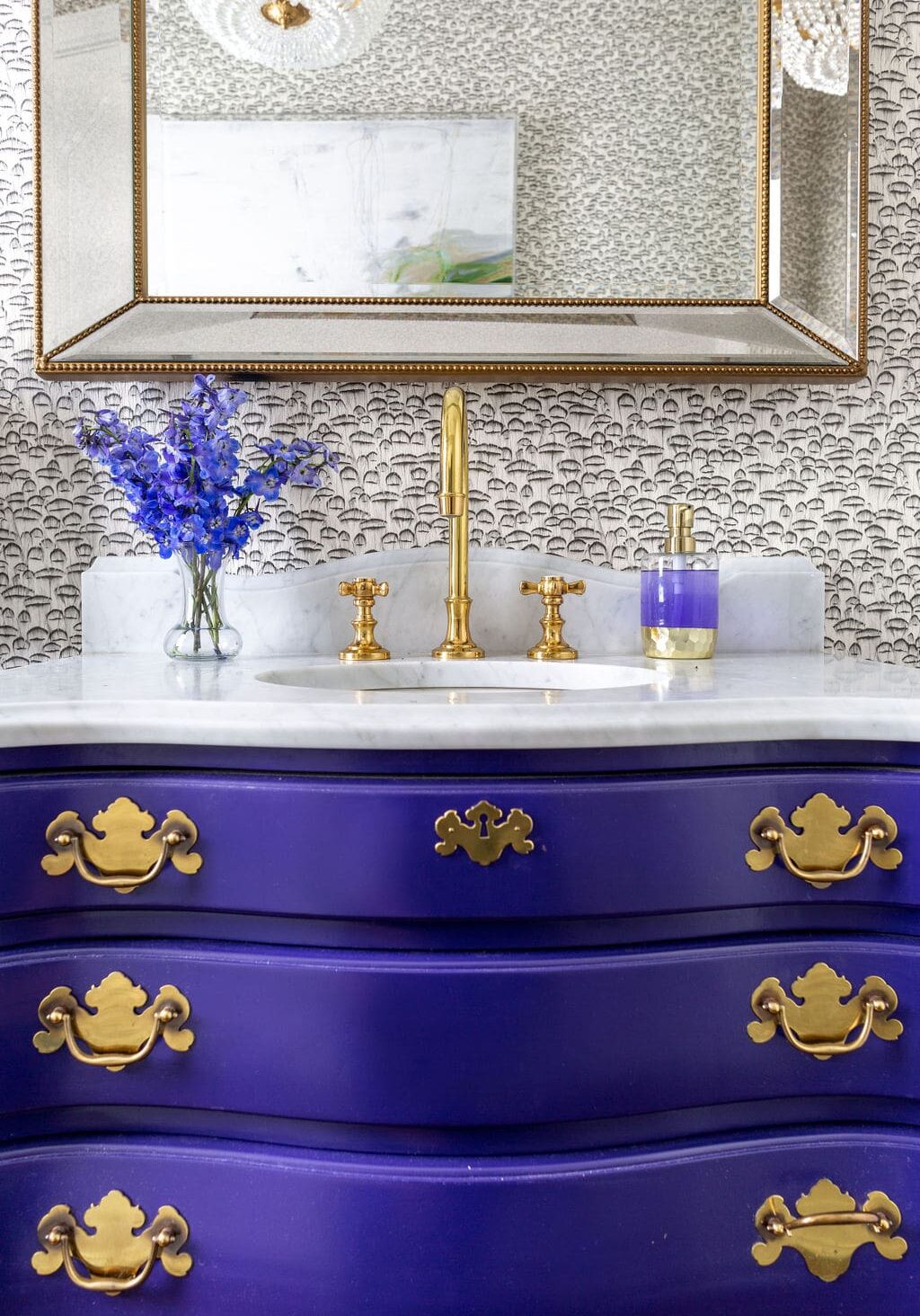A bathroom vanity with a gold faucet, marble countertop, purple drawers with gold handles, a glass soap dispenser, a small vase of purple flowers, and a large mirror above.