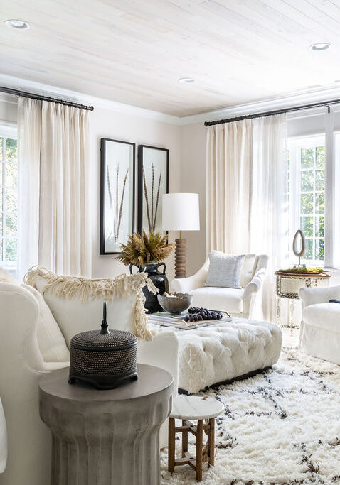 Bright living room with white walls and modern decor, featuring two armchairs, a sofa, a plush rug, coffee tables, and large windows with light curtains letting in natural light.