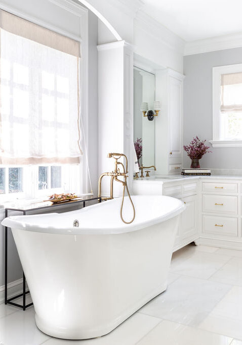 Spacious bathroom with a freestanding white bathtub, marble floors, white cabinetry, gold fixtures, and a vanity with a mirror and stool. Natural light comes through a window with a roman shade.