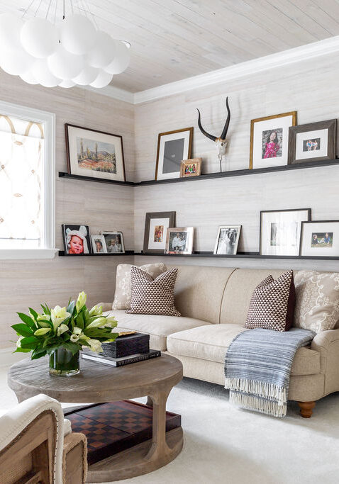 A bright living room with a beige sofa, patterned cushions, a round wooden coffee table with flowers, wall-mounted shelves holding framed photos, and antlers above the couch.