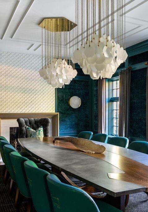 A modern dining room with a large wooden table, green upholstered chairs, geometric chandelier, dark walls, fireplace, and large windows with sheer curtains.