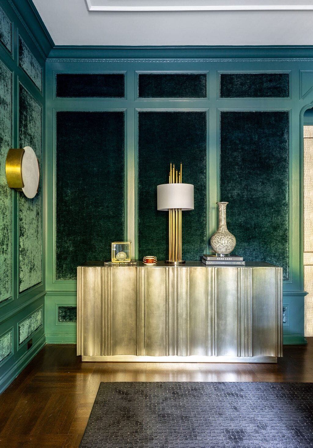 A brass console table with a modern lamp, decorative vase, and books sits against green textured wall panels in a contemporary room with a dark rug and wooden floor.