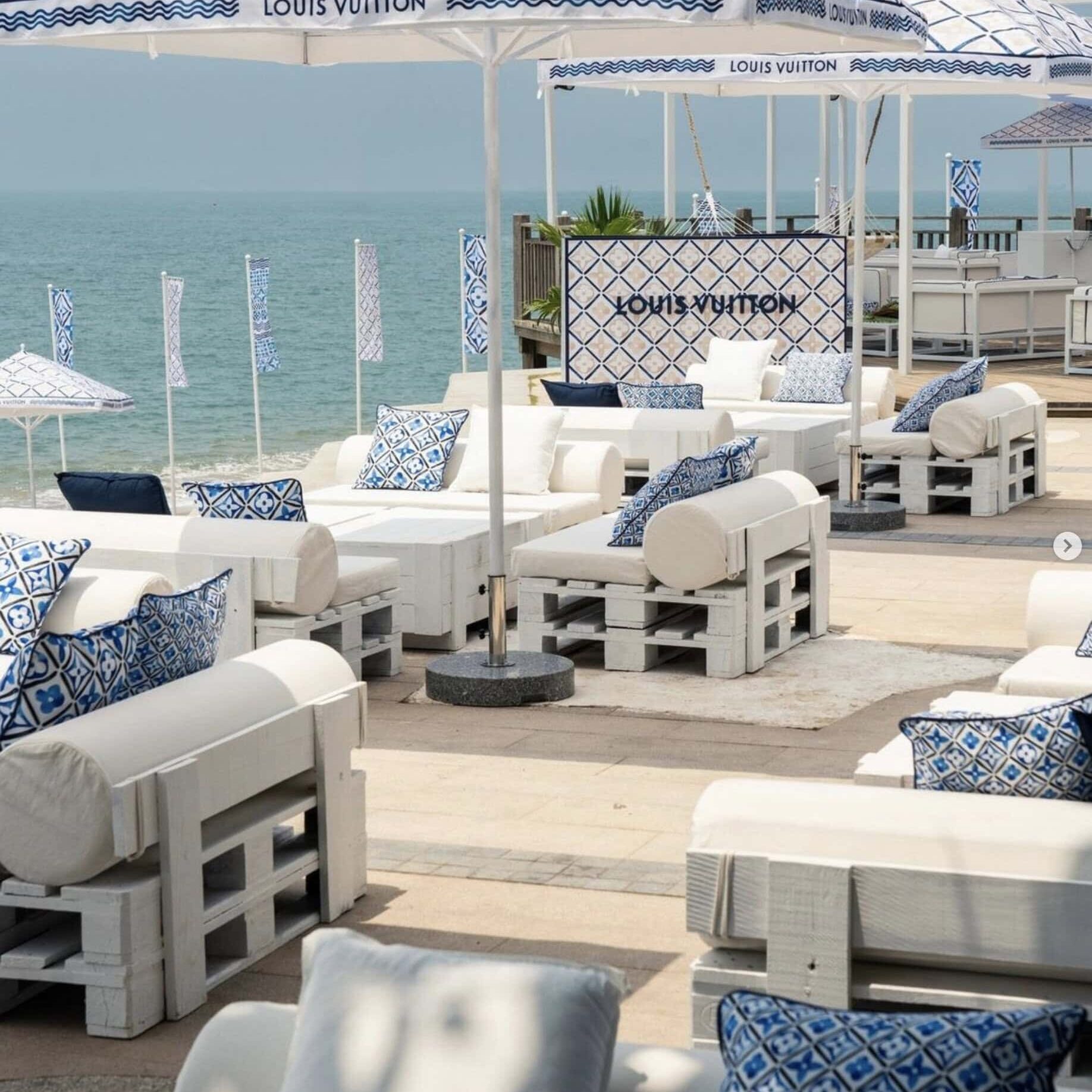 Outdoor lounge area by the sea with white wooden furniture, blue and white patterned cushions, Louis Vuitton branded umbrellas, and Crimson Design Group-inspired decor.