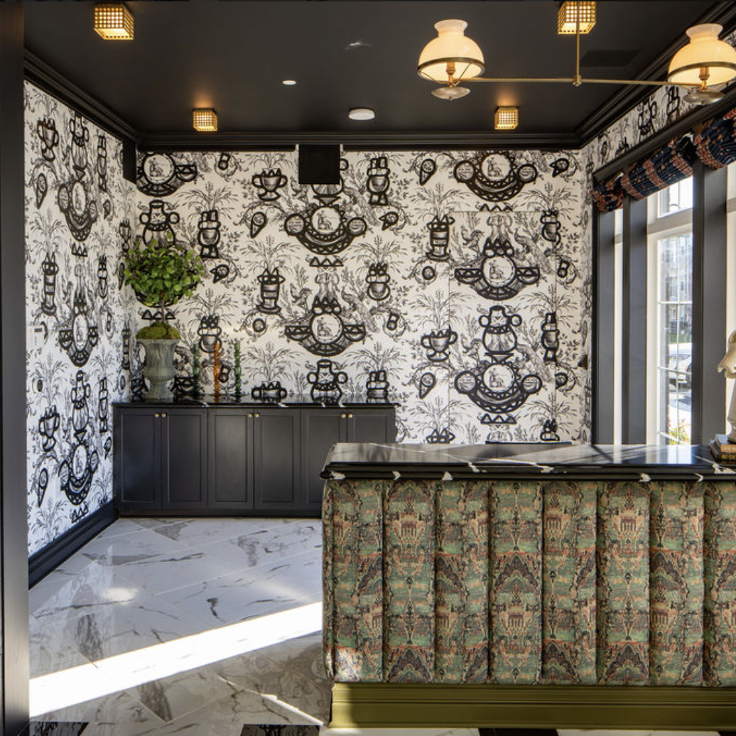 A patterned reception area by Crimson Design Group features a marble floor, black cabinets, black-and-white wallpaper, a floral arrangement, and a counter with a decorative horse head sculpture.
