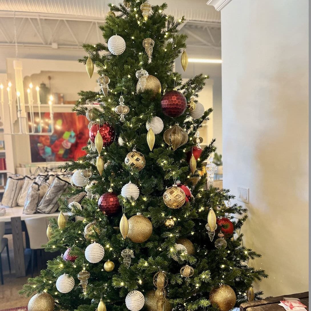 A decorated Christmas tree with gold, red, and white ornaments stands indoors near a dining area, showcasing colorful artwork in the background—a festive touch by Crimson Design Group.