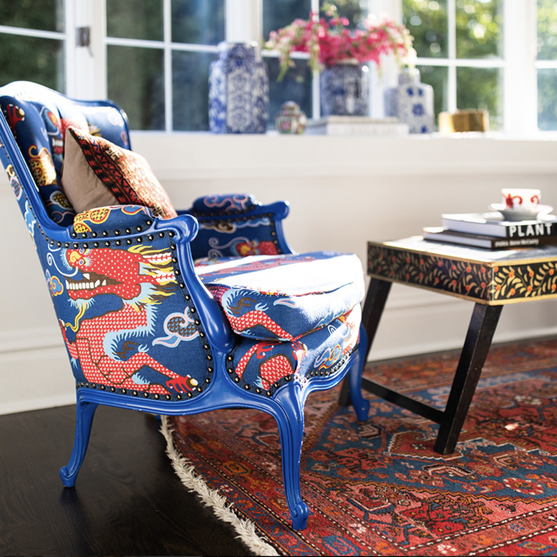 A blue upholstered armchair with a red and blue dragon pattern, styled by Crimson Design Group, sits beside a patterned coffee table on a red rug in a sunlit room with large windows and decorative vases.