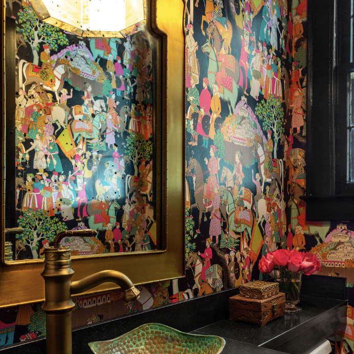 A bathroom designed by Crimson Design Group features colorful, ornate wallpaper, a gold-framed mirror, a green textured vessel sink on a black countertop, and a brass faucet. Pink flowers and decorative boxes add charm to the space.