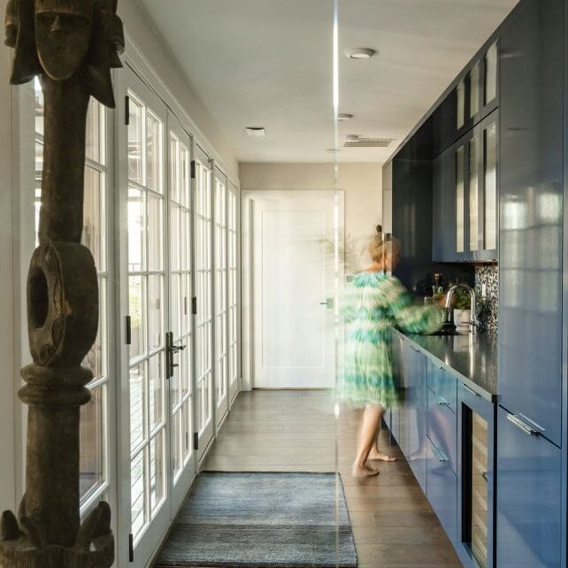 A hallway kitchen by Crimson Design Group features blue cabinets, wooden floors, and large windows; a blurred person stands at the counter preparing something.