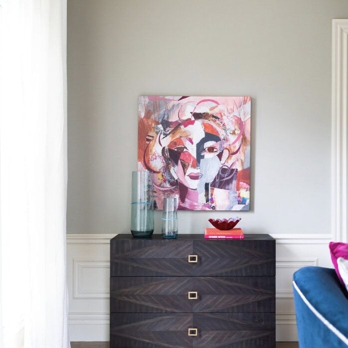 A dark wood chest with three drawers sits against a pale gray wall, topped with glass vases, a red dish, and a pink book; abstract art hangs above in this Crimson Design Group inspired space.
