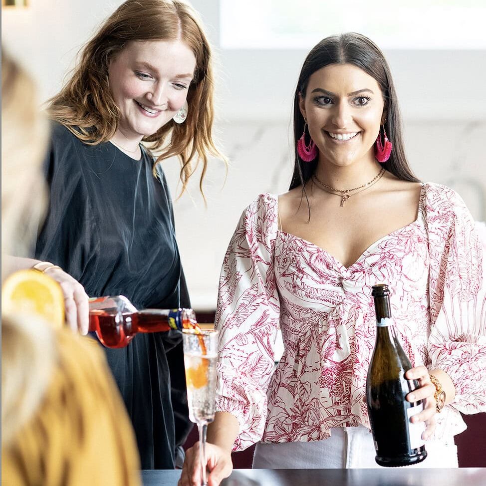 Two women stand at a table styled by Crimson Design Group, one pouring a pink drink into a glass while the other holds a bottle; a plate of cookies sits on the tastefully arranged table.