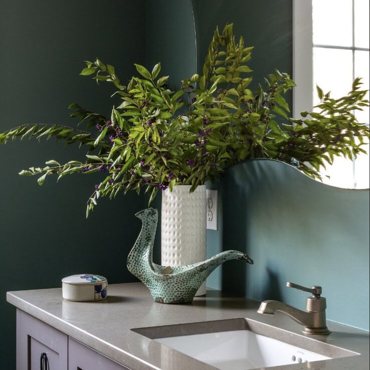 A modern bathroom by Crimson Design Group features a gray countertop, purple cabinets, a square sink, a decorative bird-shaped pitcher, a leafy vase arrangement, and a wall mirror next to a window.