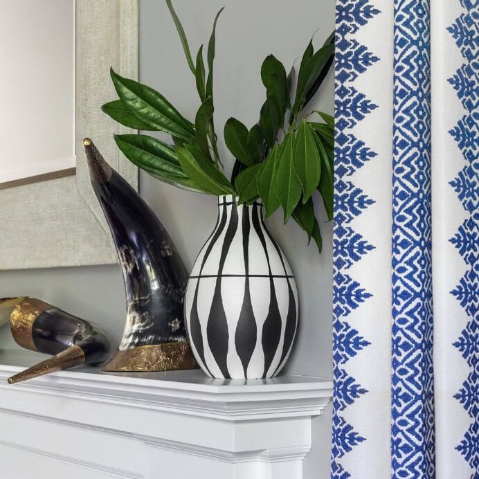 A white mantle styled by Crimson Design Group features a black-and-white striped vase with green leaves, two decorative horns, and a blue-patterned curtain in the background.