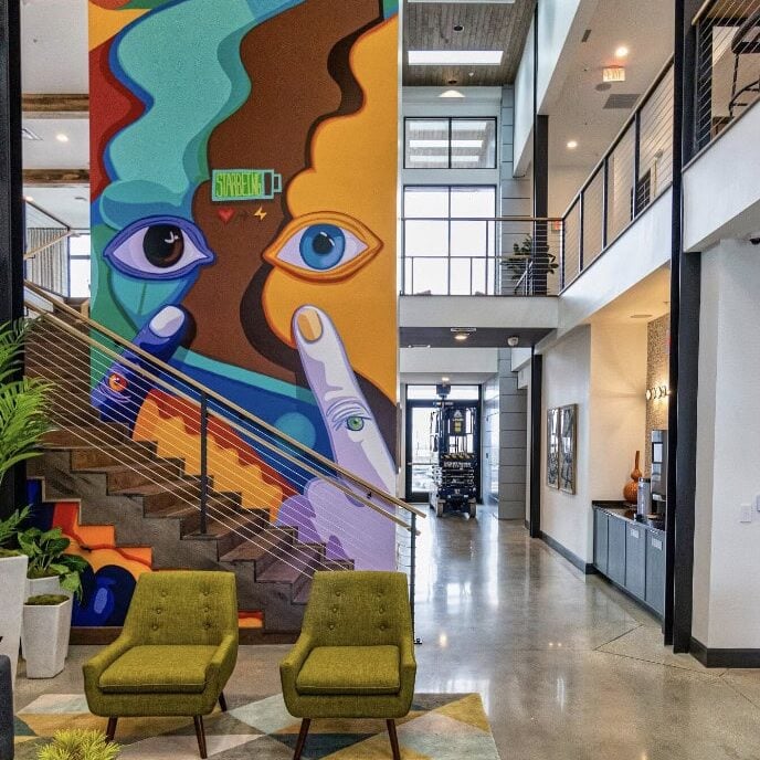 A modern lobby by Crimson Design Group with two green chairs, a geometric rug, and a colorful abstract mural featuring faces and hands beside a staircase.