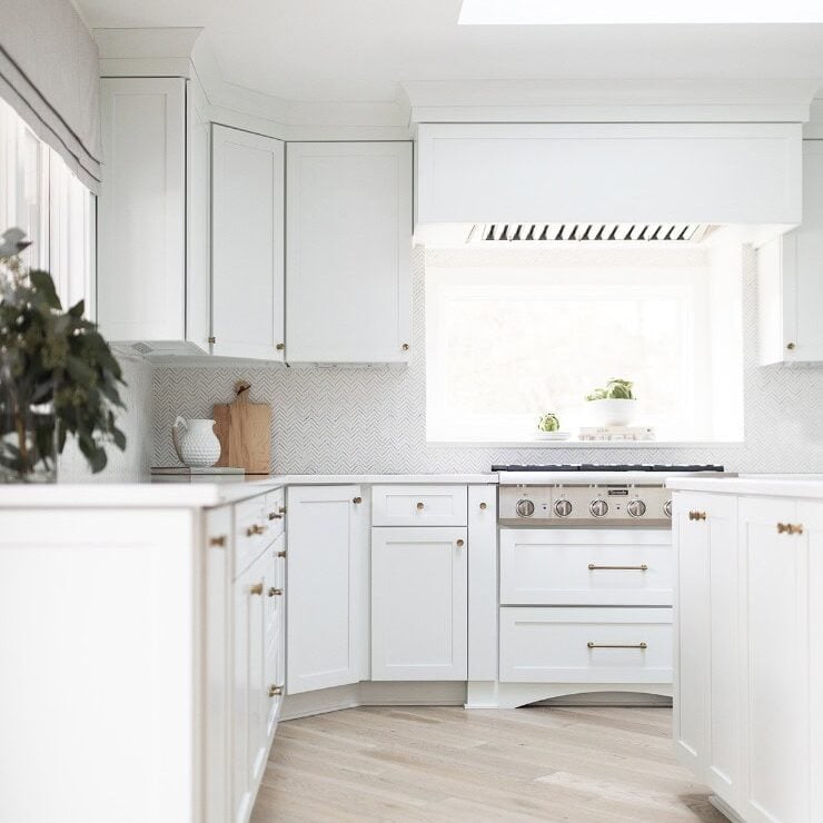 Bright, modern kitchen by Crimson Design Group with white cabinets, wood flooring, a stainless steel stove, and a large window letting in natural light.