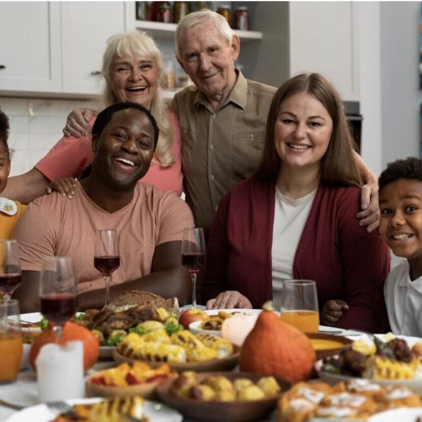 A multigenerational family of six smiles at a dining table set with various dishes, including pastries, fruit, and drinks, in a bright kitchen beautifully styled by Crimson Design Group.