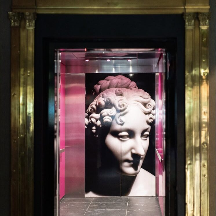 A marble statue head is depicted on the back wall of an elevator with pink side panels, framed by a gold ornate doorway and a clock above—a striking interior by Crimson Design Group.