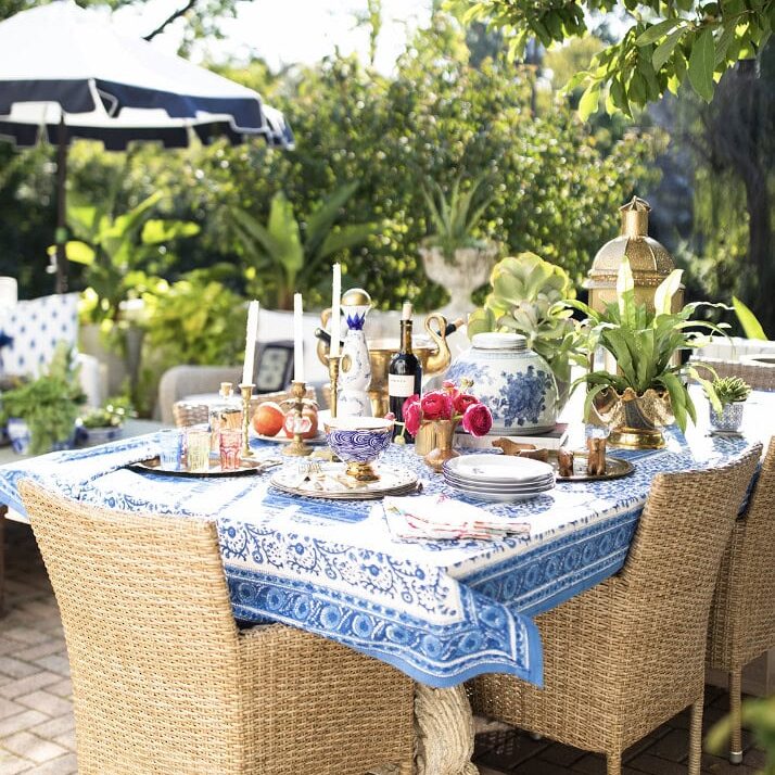 A patio table with a blue patterned tablecloth is set for a meal, featuring dishes, flowers, and decorative items styled by Crimson Design Group. Wicker chairs encircle the table, with lush greenery in the background.