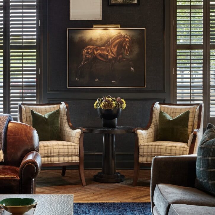 A sophisticated living room by Crimson Design Group features two plaid armchairs, a small round table with a flower arrangement, horse artwork on the wall, and large windows with wooden shutters.