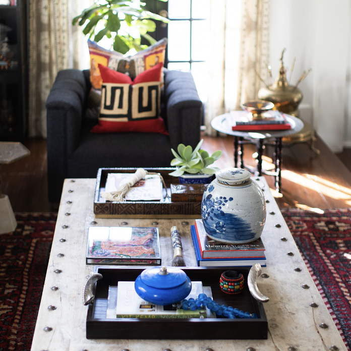 A living room by Crimson Design Group features a black armchair, red and gold pillows, a white coffee table with decorative items, books, a blue vase, and a plant by the window framed with sheer curtains.