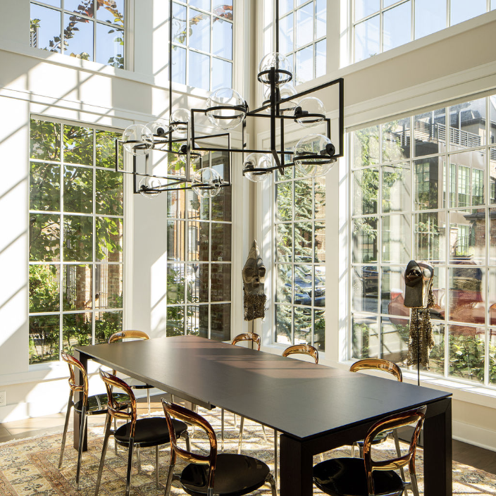 A modern dining room by Crimson Design Group features a black rectangular table, eight chairs, large windows, a patterned rug, and a geometric chandelier. Sunlight fills the space.