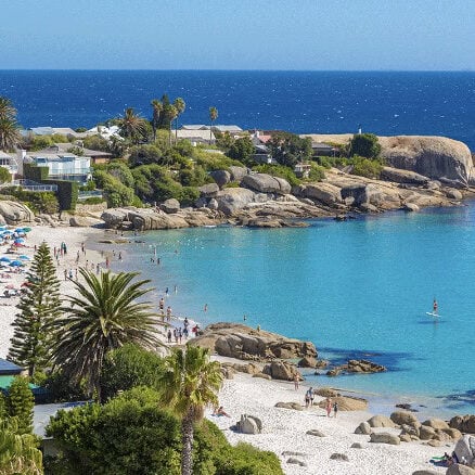 A beach with clear turquoise water, white sand, palm trees, and scattered sunbathers; houses and large boulders line the shore under a bright blue sky, echoing the vibrant aesthetic of Crimson Design Group.