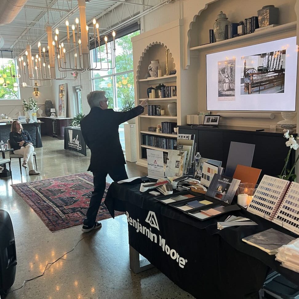 A person gestures toward a presentation screen in a modern, well-lit room with Benjamin Moore paint samples and Crimson Design Group color swatches displayed on a table in the foreground.