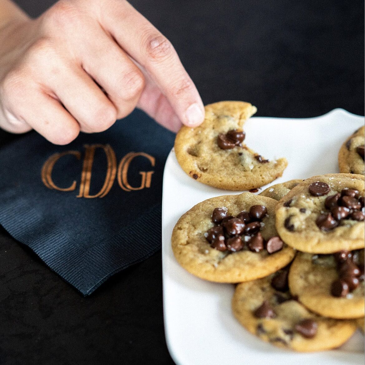 A hand reaches for a chocolate chip cookie with a bite taken out, next to a plate of cookies and a black napkin featuring gold "CDG" lettering—a nod to the sophisticated style of Crimson Design Group.