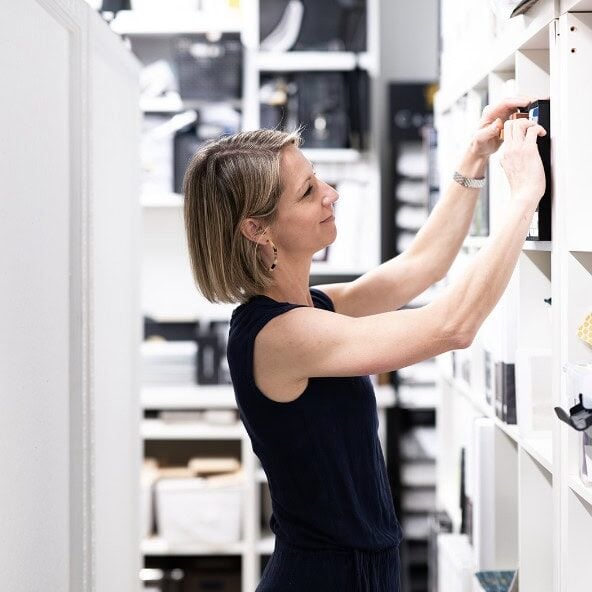 A woman in a sleeveless black outfit organizes items on white shelves in a neatly arranged storage room, reflecting the sophisticated touch of Crimson Design Group.