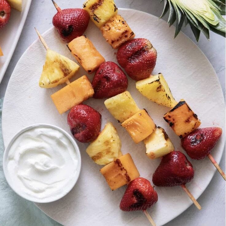 Three skewers with grilled strawberries, pineapple, and cantaloupe on a white plate next to a small bowl of yogurt and a slice of pineapple, styled in true Crimson Design Group fashion.