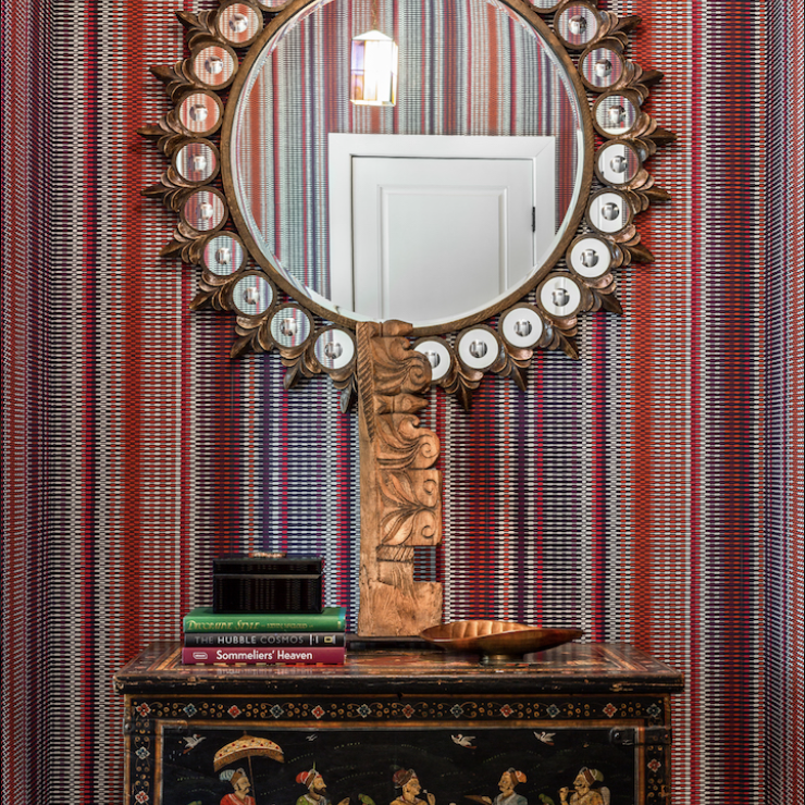 Round mirror with decorative sunburst frame above a chest featuring painted figures, set against red-striped wallpaper; books and a sculptural lamp accent the chest—an eclectic touch by Crimson Design Group.