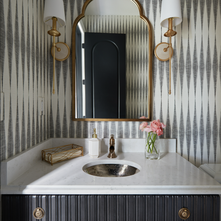 A bathroom vanity by Crimson Design Group features a marble countertop, gold fixtures, an arched mirror, two wall sconces, a vase of pink flowers, and geometric wallpaper.