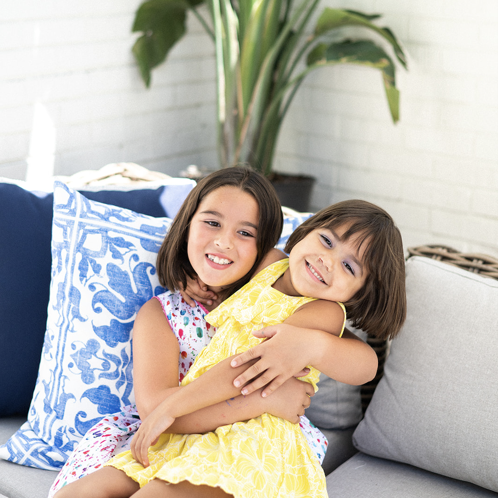 Two young girls sit on a light-colored couch; the older girl in a patterned dress holds the younger in yellow. Stylish decorative pillows and greenery, signature touches of Crimson Design Group, complete the inviting background.