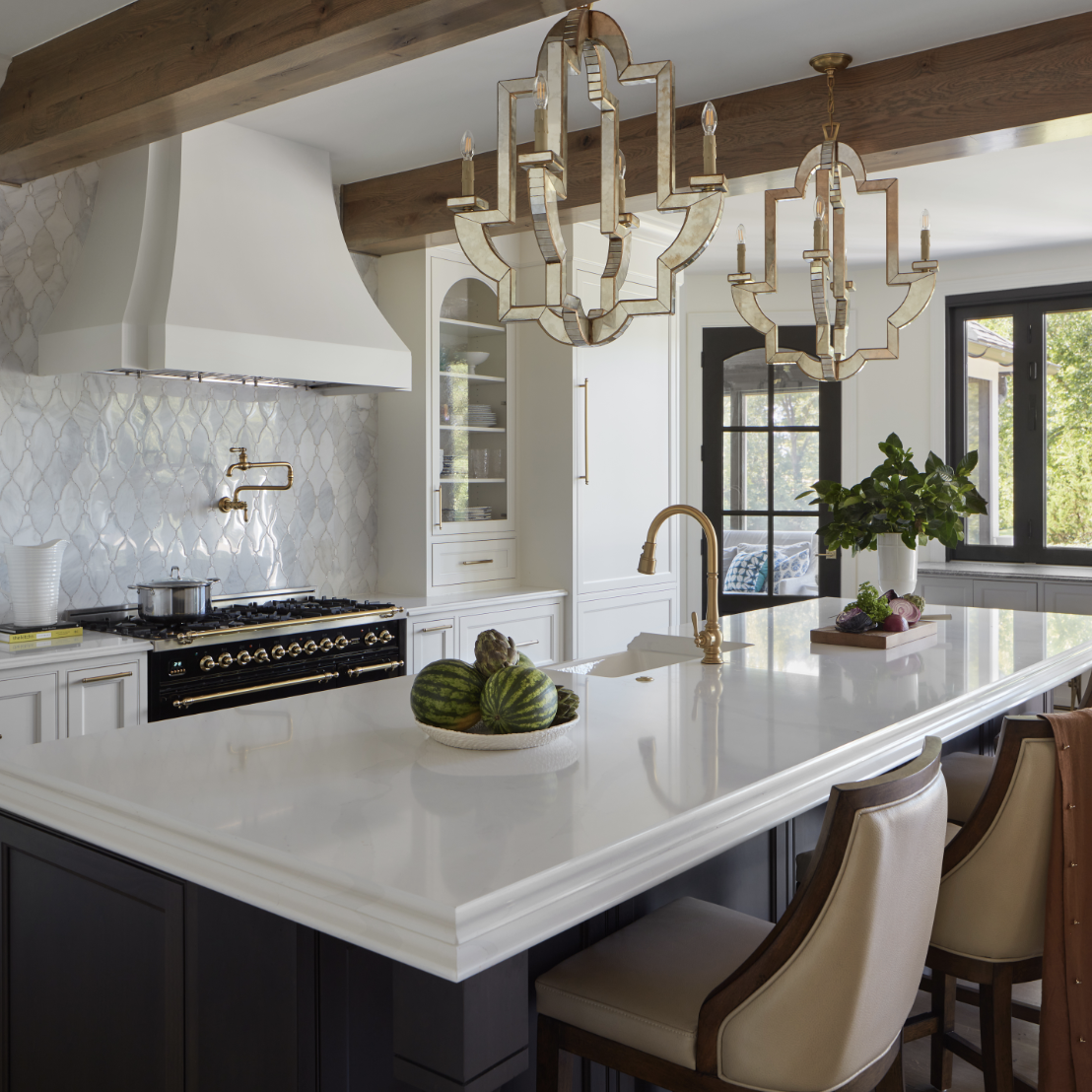 A modern kitchen by Crimson Design Group features a large white island, four chairs, gold fixtures, a black stove, white cabinetry, and two geometric pendant lights. Fruit and plants add fresh accents to the counter.