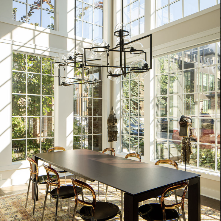 A modern dining room by Crimson Design Group features large grid windows, a black rectangular table, eight chairs, a geometric chandelier, and a patterned rug, all beautifully filled with natural sunlight.