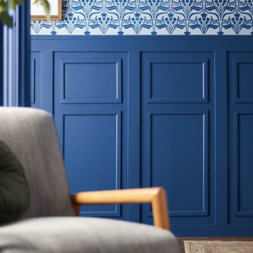 A close-up of a living room corner designed by Crimson Design Group, with a gray sofa featuring a wooden armrest, blue paneled walls, and patterned blue-and-white wallpaper above.