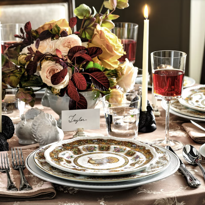 Elegant table setting by Crimson Design Group with ornate plates, silverware, a wine glass, water glass, floral centerpiece, lit candle, place card labeled "Taylor," and decorative napkin on a patterned tablecloth.