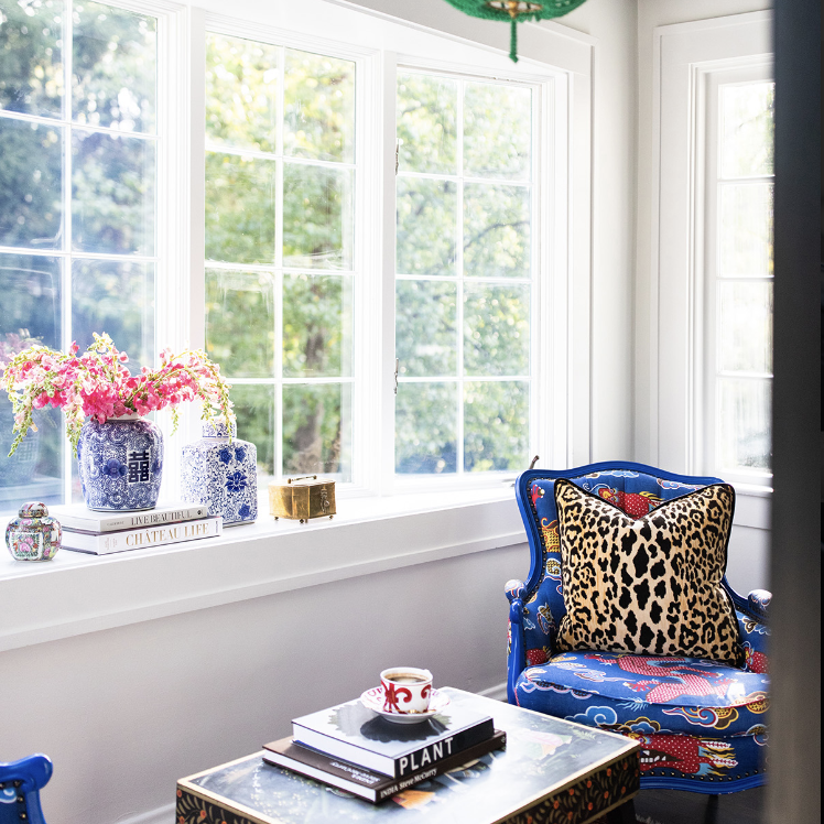 A bright sitting area by Crimson Design Group features blue patterned armchairs, a coffee table with books and a mug, a green pendant light, and large windows overlooking lush greenery.