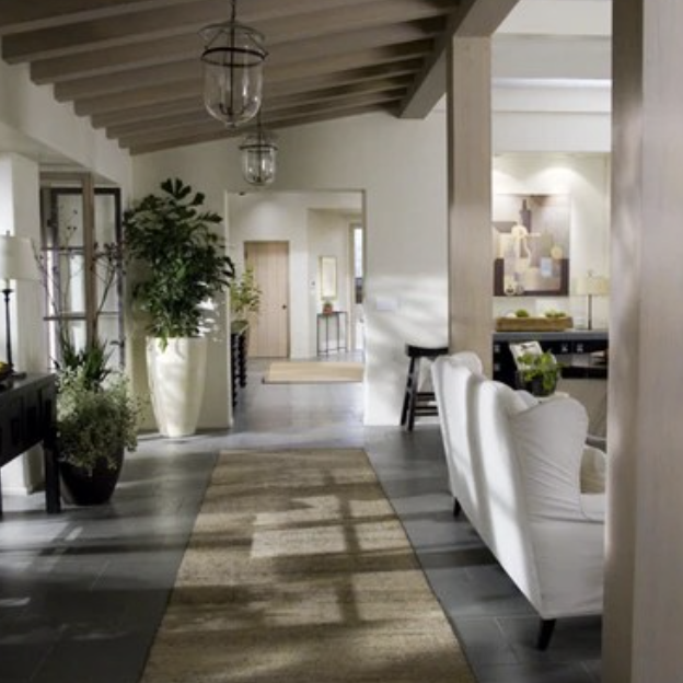 A modern living room by Crimson Design Group features white walls, exposed wooden beams, indoor plants, white sofas, a jute rug, and large windows flooding the space with natural light.
