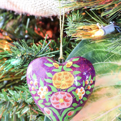 A heart-shaped ornament with colorful floral designs by Crimson Design Group hangs on a Christmas tree, surrounded by green branches and holiday lights.