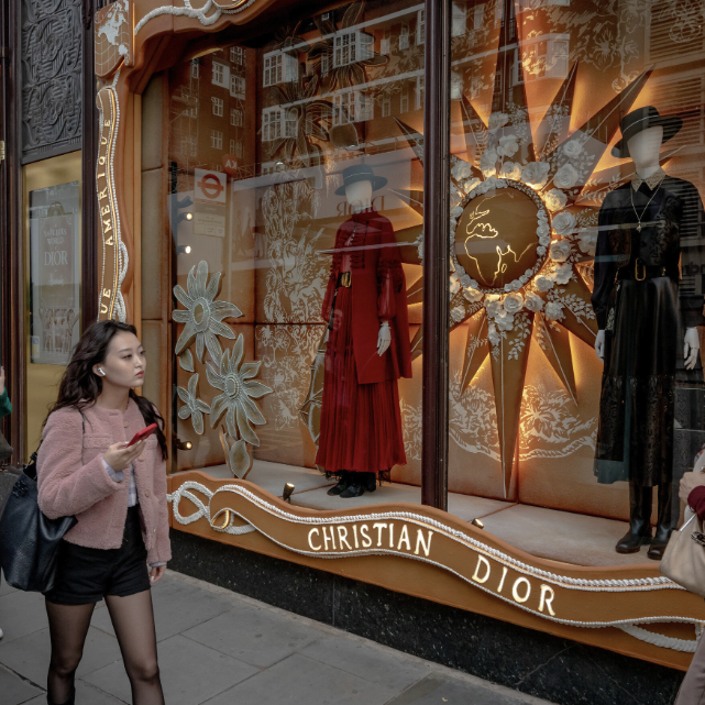 A woman walks past a Christian Dior storefront, where mannequins in elegant dresses stand before a decorative gold and floral window display inspired by Crimson Design Group.